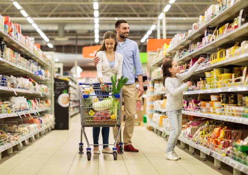 A family of three browses the aisles of a grocery store, with the parents pushing the cart while their child raises an arm to a shelf.